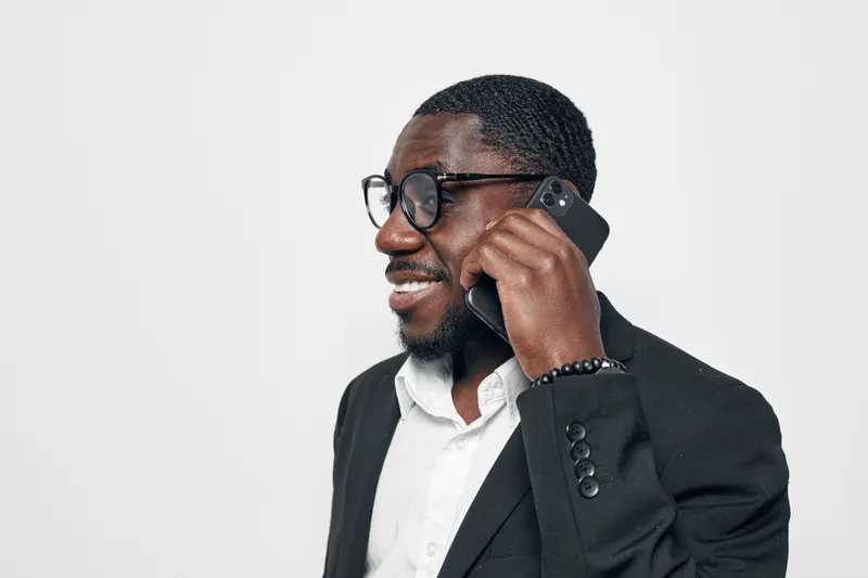 Black African man with dark skin wearing glasses and black suit talking on smartphone smiling isolated on white background. Business professional, communication and technology concept.