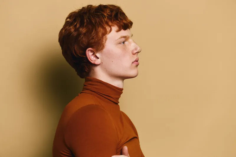 Profile portrait of young man with red hair wearing brown turtleneck sweater looking forward against plain yellow background, side view with serious expression and casual style.