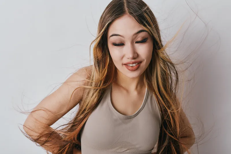 Young woman with long hair wearing beige top posing against white background with closed eyes and slight smile. Casual portrait with natural light and relaxed expression.