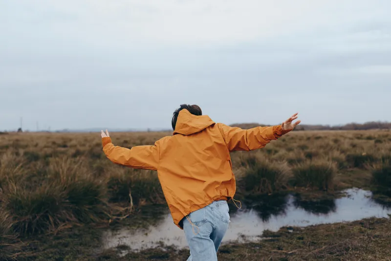 Woman in outdoor orange jacket with arms outstretched walking in grassy field by water puddle on autumn day, nature lifestyle, overcast sky, soft light, scenery, calm mood, in wind, today