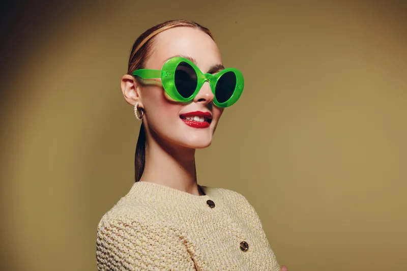 Stylish woman wearing large green sunglasses and a beige knitted sweater smiles confidently against a plain background. Concept of fashion, accessories, and modern style.