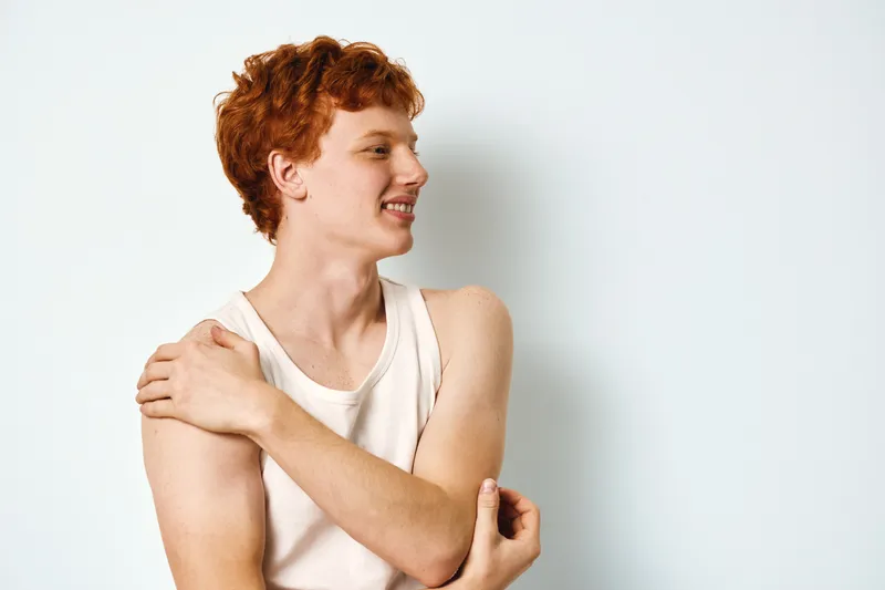Young man with red hair wearing white sleeveless shirt touching shoulder against white background for skincare or health concept with casual pose and smiling face.
