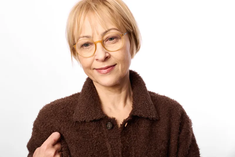Portrait of a senior woman with short blonde hair and glasses, wearing a brown coat, smiling at the camera in a studio with white background, soft light.