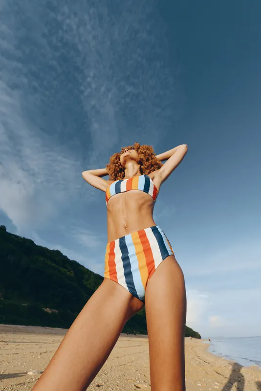 Woman in striped swimsuit on sandy seaside beach near ocean in summer sunlight, arms behind head, relaxation pose, clear blue sky, warm coastal breeze, sunlit sandy shore, vacation mood