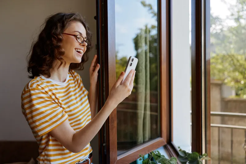 A woman stands by an open window, wearing a striped tee, smiling as she checks her phone. Natural light fills a bright, cozy home space, capturing a casual, relaxed lifestyle.