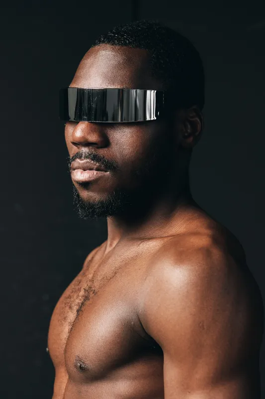 Black man with dark skin wearing futuristic reflective visor glasses in creative conceptual visual style, bare chest and confident pose on black background for modern portrait.