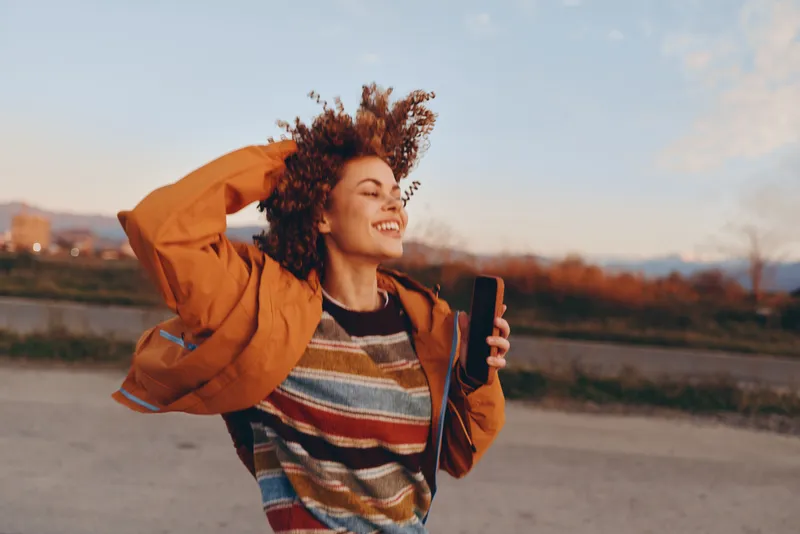 Smiling woman with curly hair, orange jacket, holding smartphone outdoors on a road at sunset, autumn lifestyle and communication, casual fashion, warm evening light, cheerful mood
