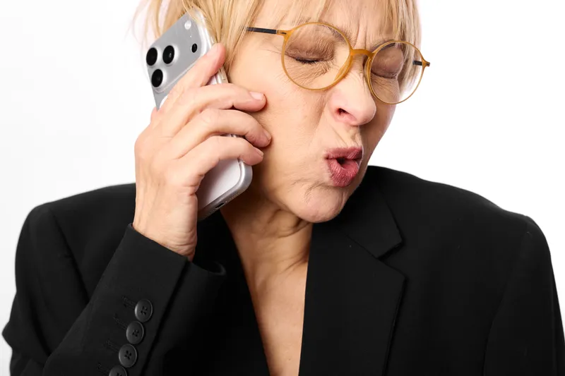Elderly woman in business suit with glasses talks on phone, surprised expression, closeup on white background, office communication concept, customer support