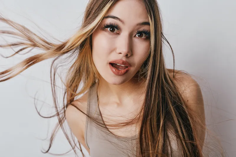 Young woman with long hair blowing in wind wearing tank top looking at camera with open mouth on white background. Beauty, fashion and portrait with natural expression and movement.