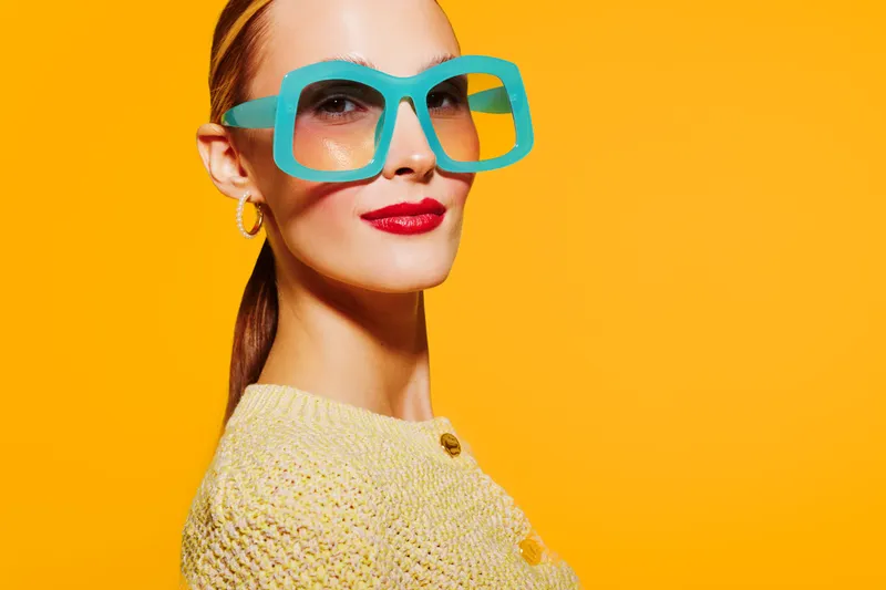Stylish woman with large turquoise glasses and red lipstick poses confidently against bright yellow background. Fashion portrait with vibrant colors and modern accessories concept.
