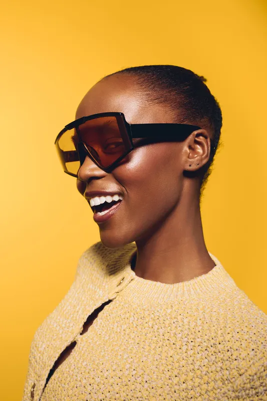 Stylish African woman with dark skin wearing large sunglasses and a knitted sweater smiles confidently against a bright yellow background. Concept of fashion, beauty, and happiness.