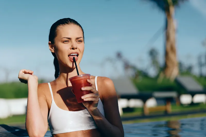 Poolside woman outdoors, hydration and summer lifestyle, drinking a refreshing beverage, sunlit moment by the pool, casual athletic vibe, vibrant colors, relaxed mood, greenery in background
