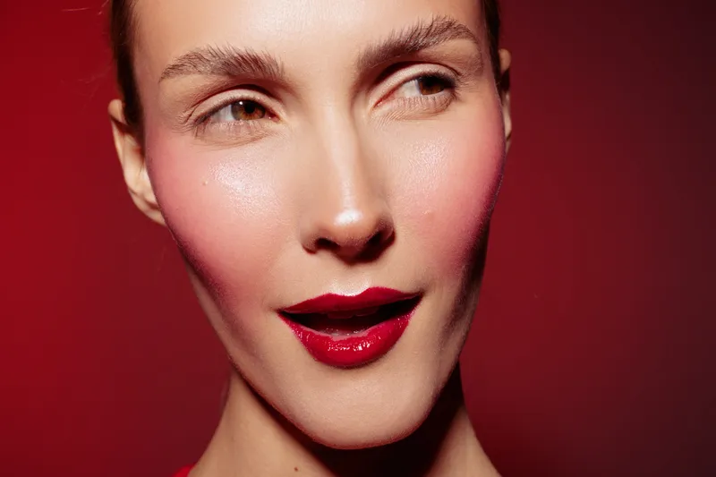 Close-up of a woman with glossy red lips and pink blush on cheeks against a red background. Concept of makeup, beauty, skincare, and fashion with studio lighting.