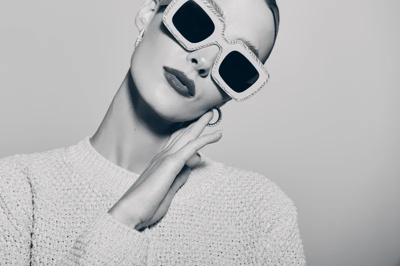 Stylish white woman with fair skin wearing oversized sunglasses and textured sweater poses against plain background. Concept of fashion, elegance, and modern style.