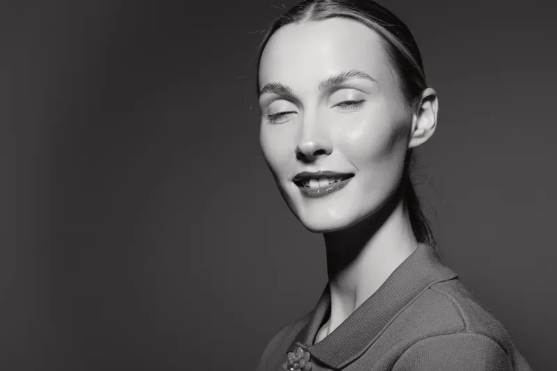 Portrait of a smiling young white woman with pale skin and closed eyes in studio. Elegant female model expresses calm and confidence, monochrome image with soft lighting.