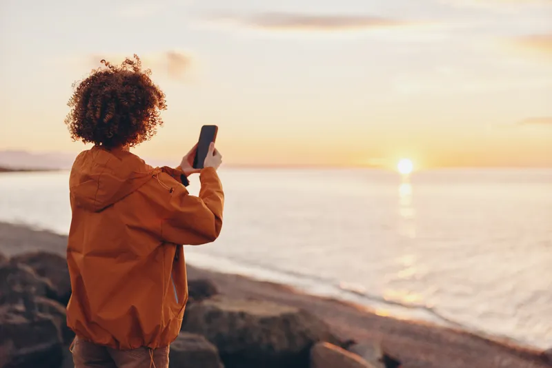 Woman with curly hair standing at the seaside during sunset, holding a smartphone and looking at the screen, casual outdoor lifestyle, warm golden light, calm ocean soft waves