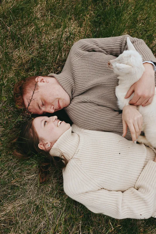 A couple lounges on green grass, cuddling a white goat kid in oversized knit sweaters, creating a relaxed countryside moment. A cozy outdoor scene that captures affection, nature, and simple farm