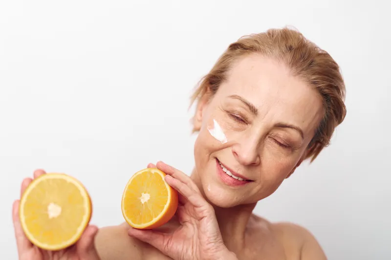 Smiling mature woman with natural skin holds two orange halves near face with cream on cheek. Concept of skincare, healthy lifestyle, natural beauty, and vitamin c benefits.
