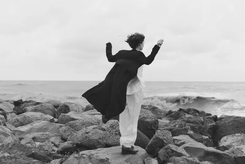 Two figures embrace on a rocky shoreline, wind catching coats as the sea meets the jagged coast, a black and white scene of movement, freedom and connection