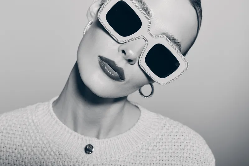 Stylish white woman with pale skin wears oversized sunglasses and knitted sweater in black and white studio portrait concept for fashion and elegance.