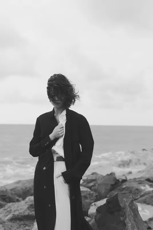 Black and white photo of a woman in a long coat standing on a rocky shoreline, wind sweeping her hair, one hand at the lapel, ocean calm in the background, moody fashion and solitary moment