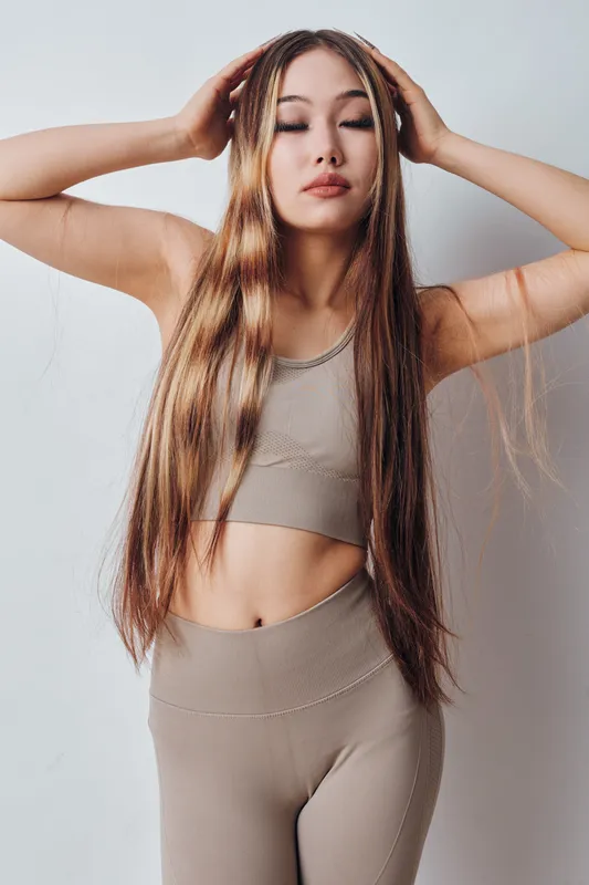 Young woman with long hair wearing beige sportswear standing against white wall with closed eyes and hands touching head and hair in natural light.