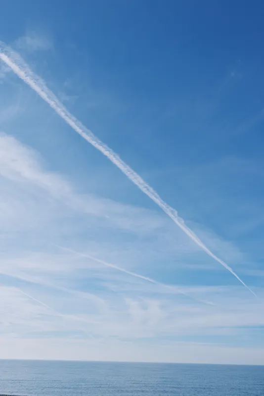 Blue sky over calm ocean with a distant horizon and a white contrail streak, gentle clouds, serene seascape conveying tranquility and expansive outdoor scenery.