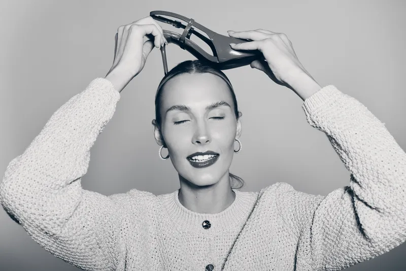 Young white woman with light skin and slicked hair holds a high heel shoe above her head, eyes closed, wearing a textured sweater and hoop earrings in a studio setting.