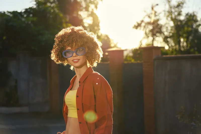 A young woman with curly hair wearing sunglasses and a bright yellow crop top under a red jacket stands in a sunlit street, conveying a casual, confident lifestyle moment on a warm afternoon