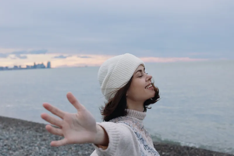 Woman on a pebble beach reaches toward the camera, smiling, wearing a knit sweater and beanie, with calm sea and soft clouds behind.