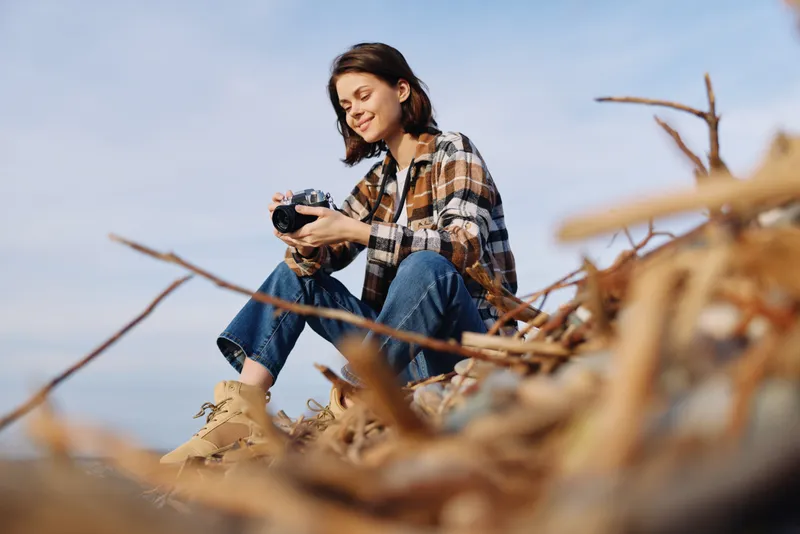 Woman photographer in a plaid shirt sits on sandy dunes by the coast, holding a camera and smiling at the view in a bright outdoor scene today.