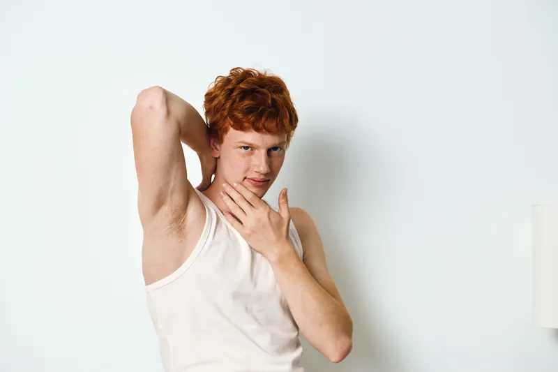 Young man with red hair wearing white tank top posing with hand under chin and other raised showing armpit in bright room with white wall background.