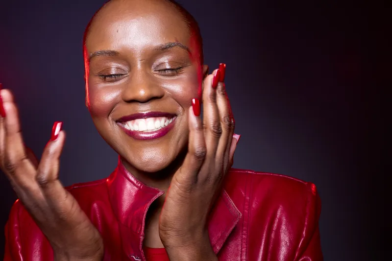 Happy dark skinned African woman with red jacket and manicured nails smiles with closed eyes on dark background. Concept of beauty, joy, confidence, self care, and fashion portrait.