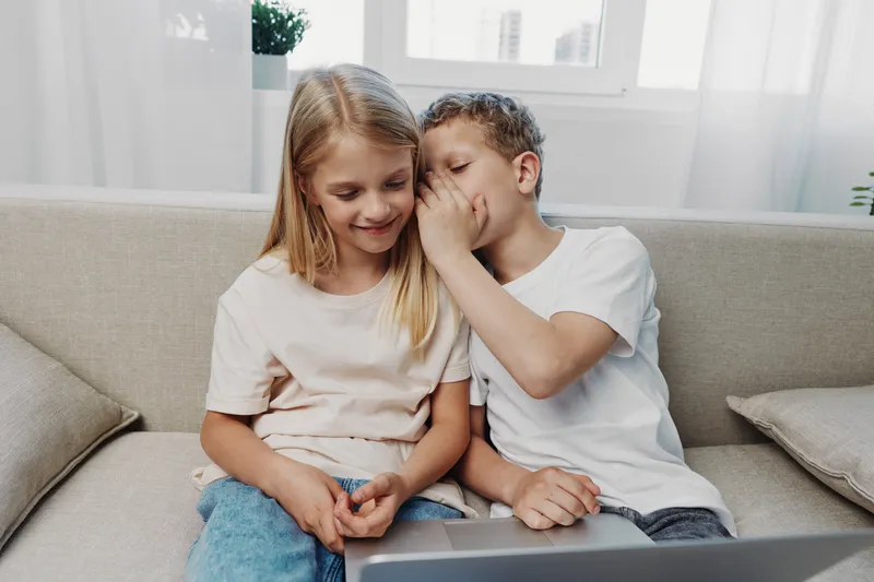 A young boy whispers a secret to a smiling girl sitting next to him on a sofa in a bright living room. The children share a moment of friendship and playful communication.