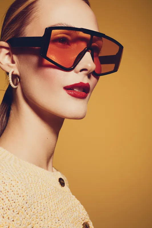 Stylish woman with oversized orange sunglasses and red lipstick poses against yellow background. Fashion portrait with modern accessories and confident expression.