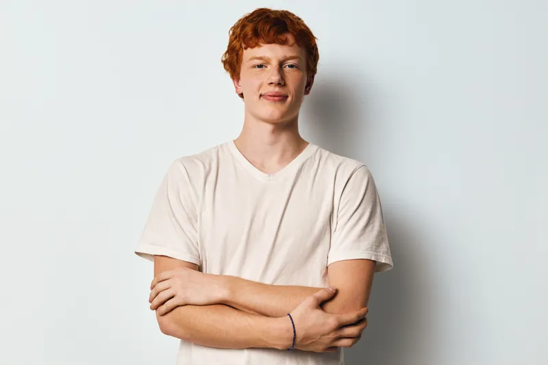 Young man with red hair and casual white t shirt stands confidently with arms crossed against plain light background in studio. Portrait with natural expression and relaxed posture.