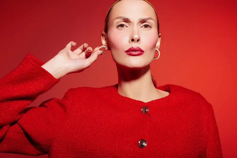 Confident woman in red sweater poses against red background with hand near ear. Studio portrait with bold makeup and hoop earrings. Concept of fashion, style, and self-expression.