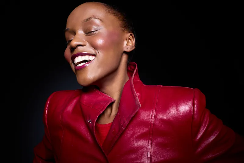 Happy African woman with dark skin laughs joyfully wearing a red leather jacket. Studio portrait on black background. Concept of fashion, beauty, confidence, and positive mood.