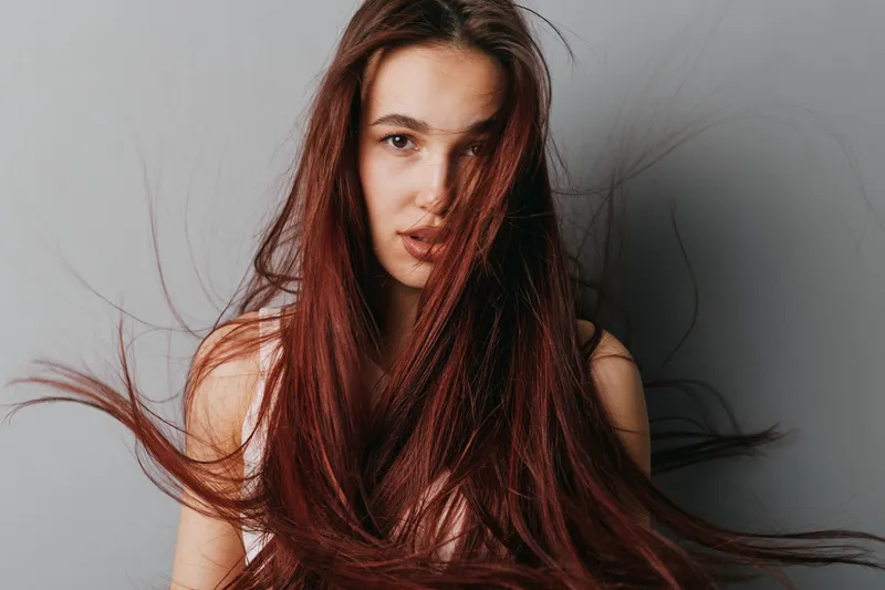 Young woman with long red hair blowing in the wind, looking at camera with serious expression in studio. Portrait of female model with natural makeup and loose hair strands.