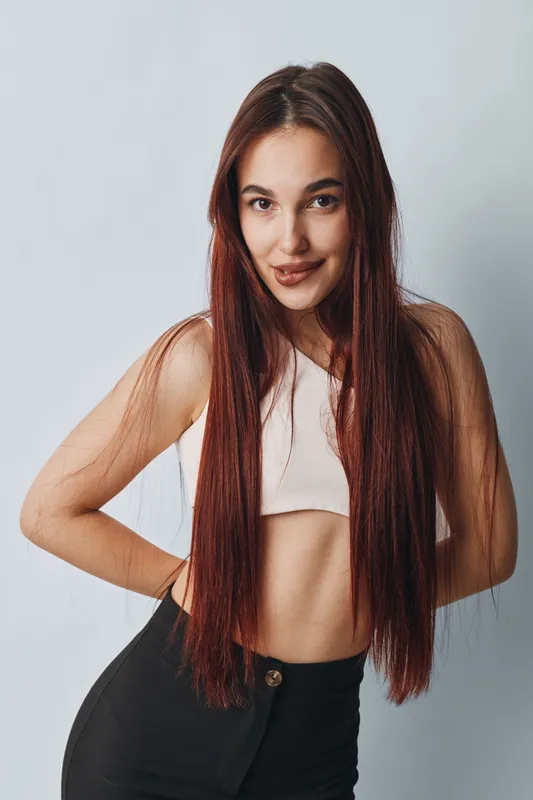 Young woman with long straight hair posing with hands on hips in casual outfit. Studio portrait with confident smile and natural makeup on light background.
