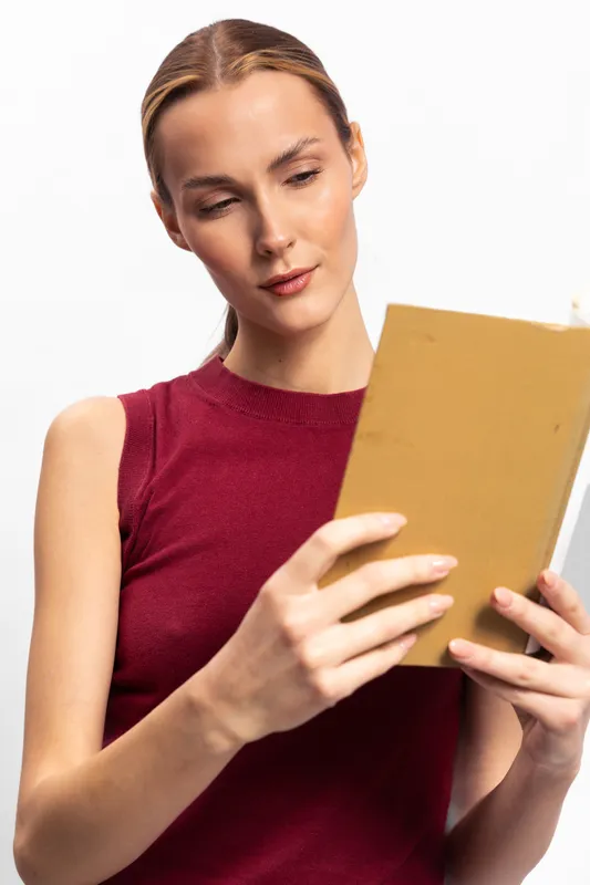 Young woman in red sleeveless top reads a book with focused expression. Studio shot on white background. Concept of education, learning, and concentration.