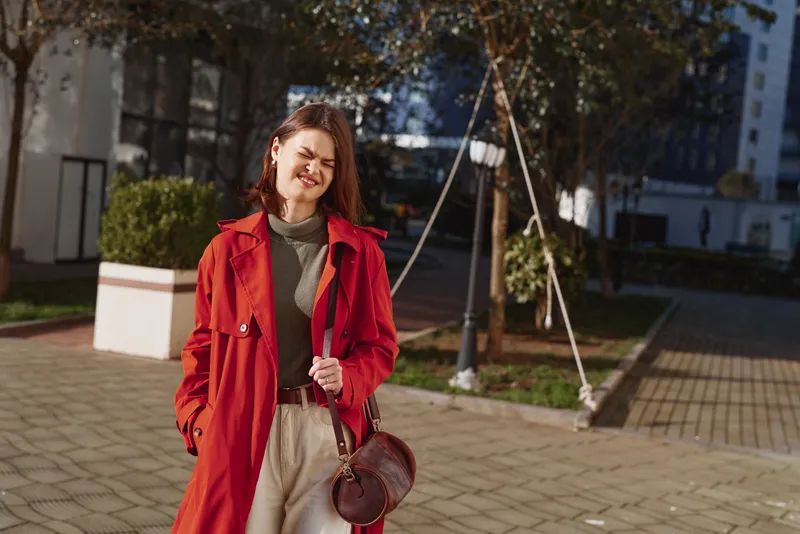 Redcoat wearing woman smiles outdoors on a sunny street, casual fashion, handbag, urban setting, confident pose, autumn light, relaxed mood, lifestyle portrait, contemporary style