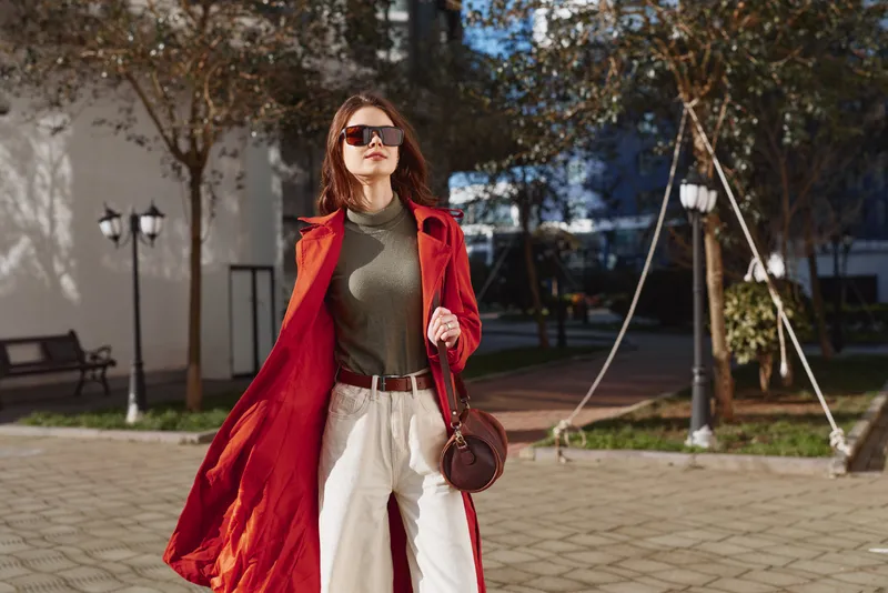 Stylish woman wearing a red coat and sunglasses walking outdoors on a sunny day. Urban background with trees and pavement. Concept of fashion, confidence, and autumn style.