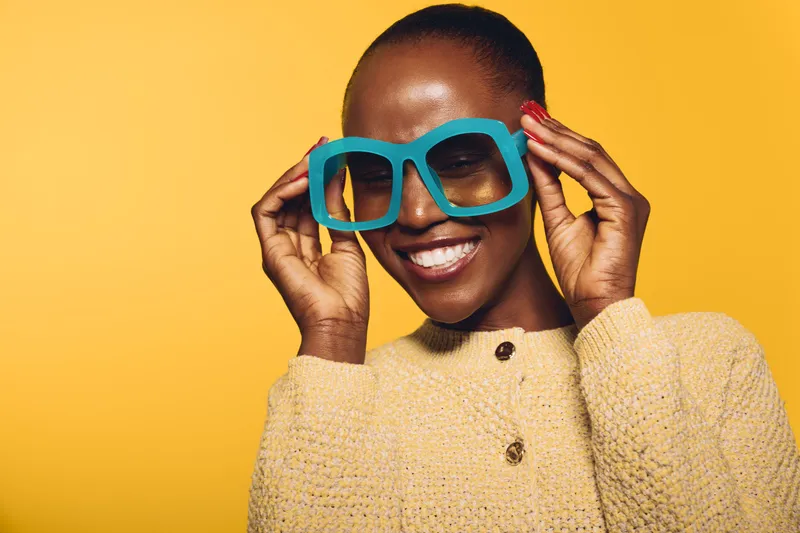 Smiling African woman with dark skin wears oversized blue glasses and a beige sweater. Bright yellow background highlights her joyful expression and stylish casual look. Concept of fashion and