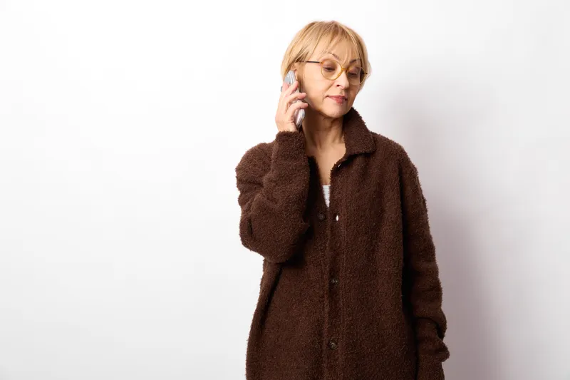 Senior woman with blond hair holding a smartphone near her ear, portrait on white background, communication by phone, calm relaxed expression, indoor studio portrait