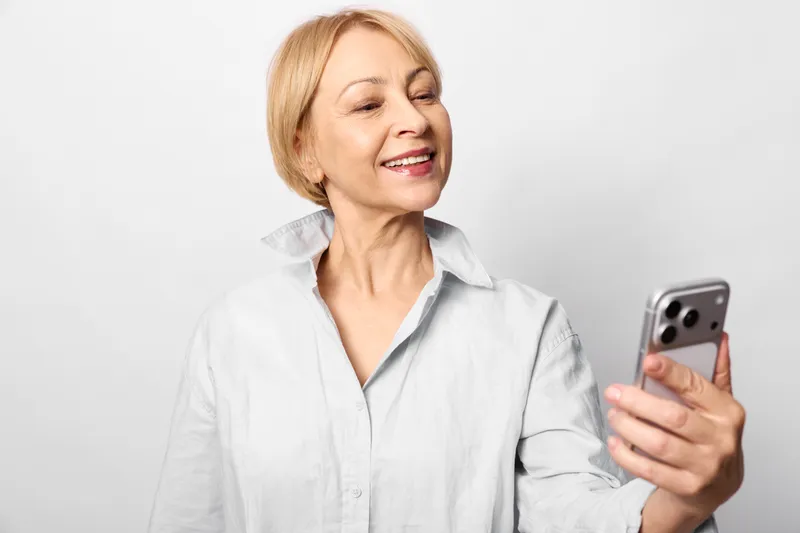 Elderly woman smiling, holding smartphone, casual portrait indoors, white background, friendly senior using digital communication, remote video call, relaxed mood