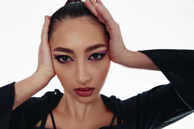Close-up of a young woman with bold colorful eye makeup and glossy lips adjusting her hair against a white background. Concept for creative beauty, makeup art, and fashion portraits.