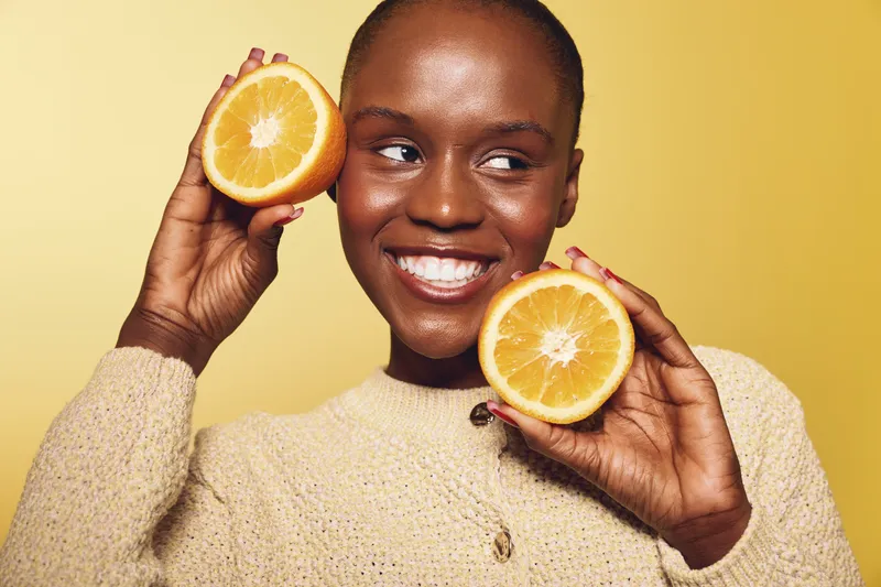 Smiling African woman with dark skin holds two orange halves near face against yellow background. Concept of healthy lifestyle, vitamin C, natural beauty, and fresh fruit wellness.