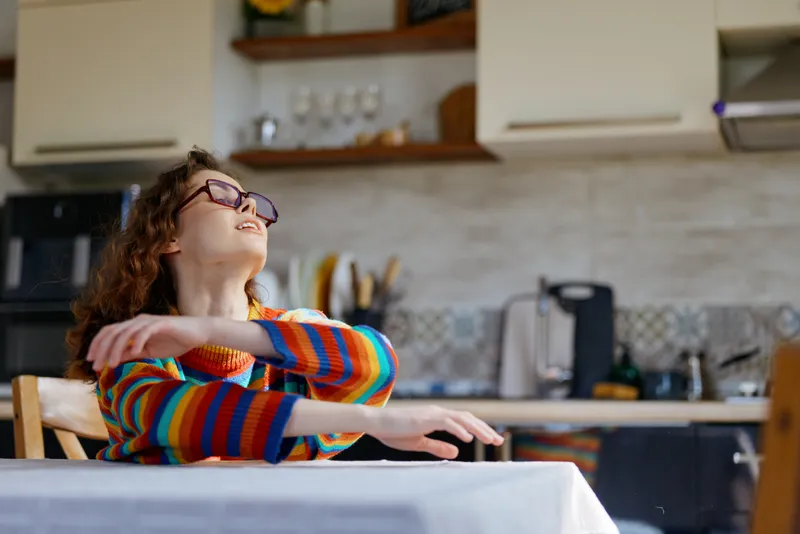 Woman with a colorful striped sweater smiles and leans back in a bright kitchen, enjoying a carefree moment at the table, capturing a warm home lifestyle