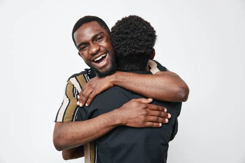 Two black men hugging each other with happy expressions on white background. African ethnicity, dark skin, friendship, connection, joy, casual clothing, positive emotions, studio portrait.